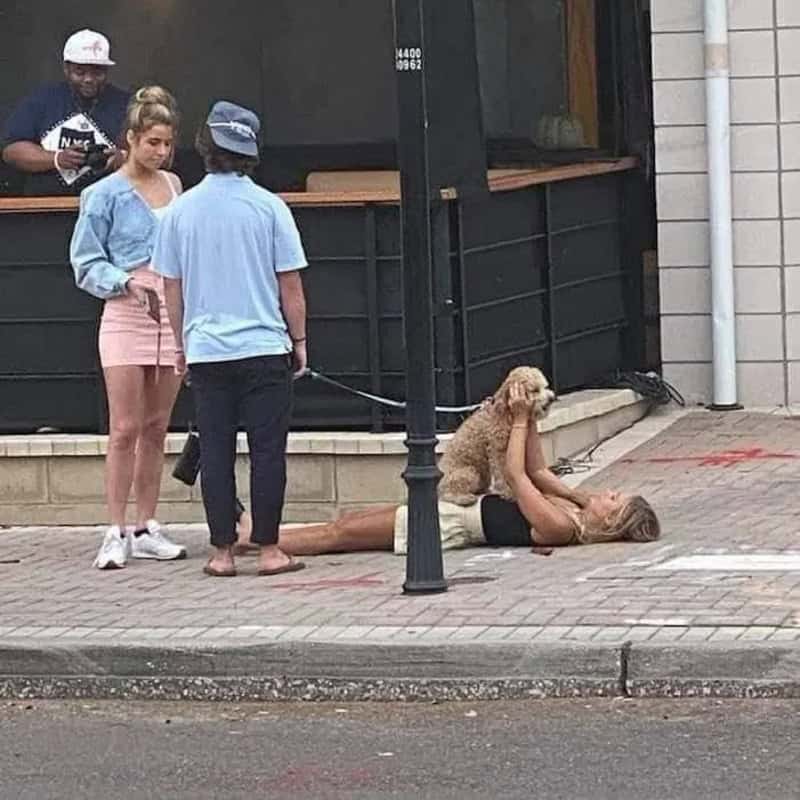 Woman lying on the sidewalk with a dog on her face, people watching.