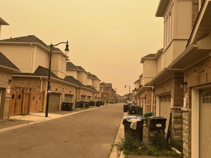 Empty suburban street with houses and trash bins, under a yellowish sky.