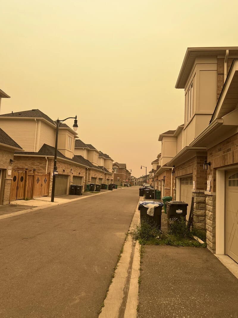 Empty suburban street with houses and trash bins, under a yellowish sky.