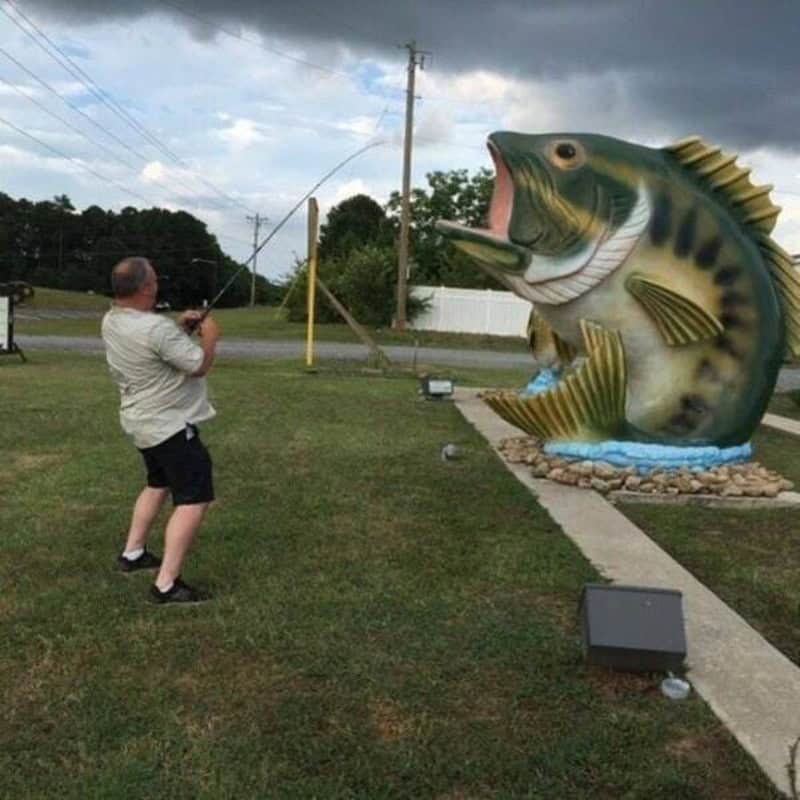 Man fishing at a giant fish statue in a park during cloudy weather.