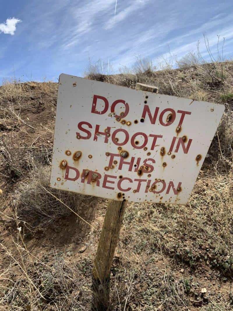 Rusty sign says Do Not Shoot In This Direction, tilted on a post in a dry area.