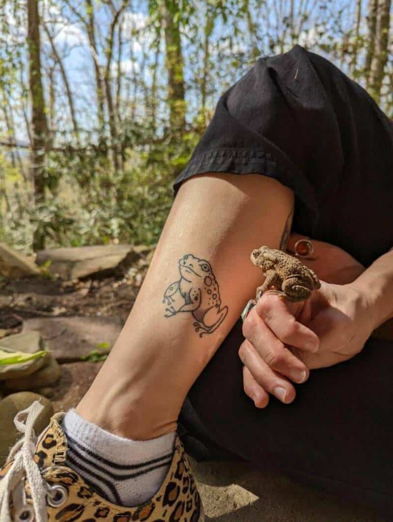 Person holding a small lizard with a tattoo of a frog on their arm.