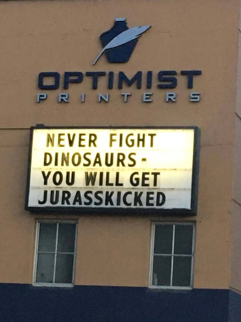 Sign reads 'NEVER FIGHT DINOSAURS - YOU WILL GET JURASSKICKED' at Optimist Printers.