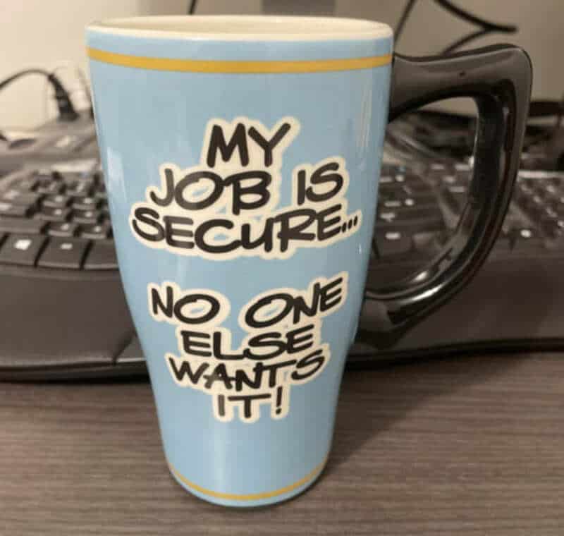 Coffee mug with the text "My job is secure... no one else wants it!" on a desk.
