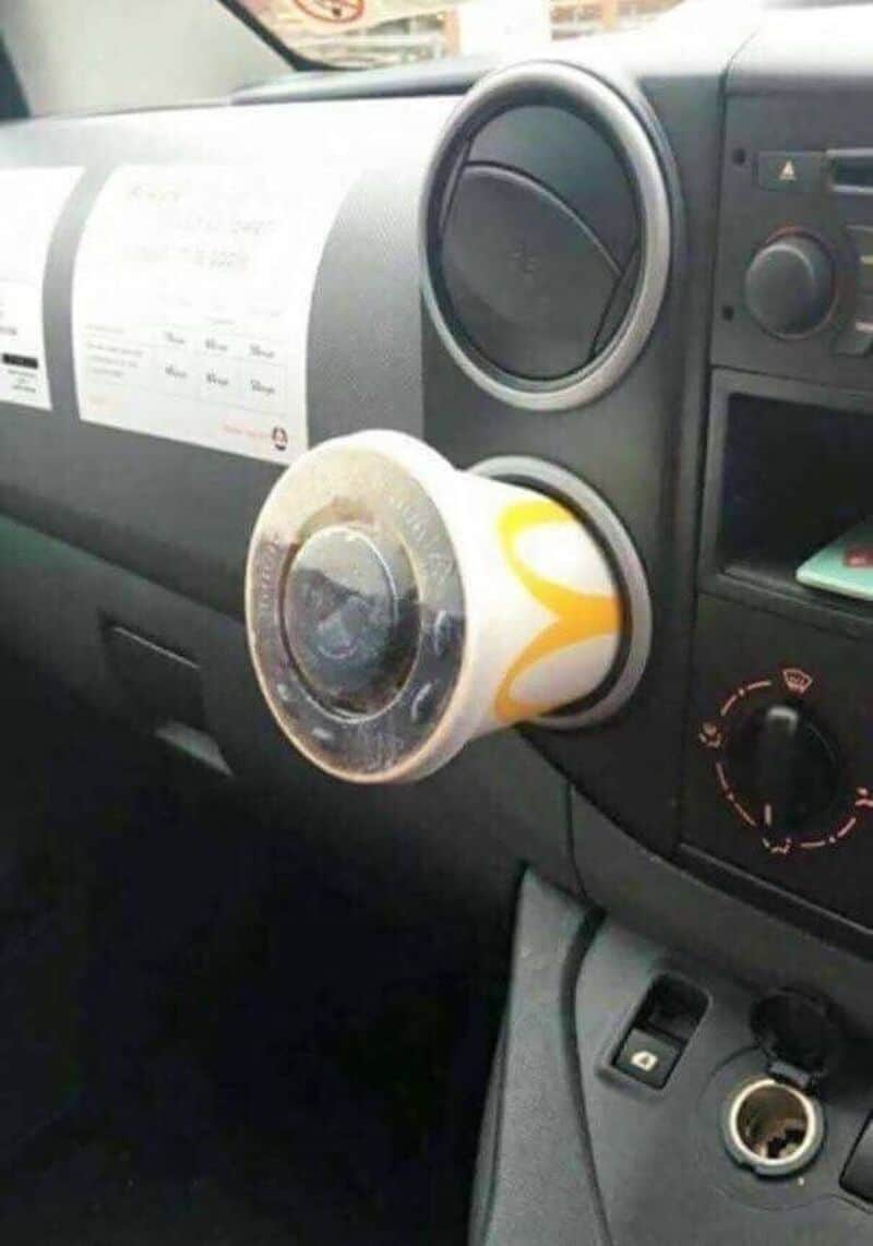 A coffee cup attached to a car's air vent, with a receipt on the dashboard.