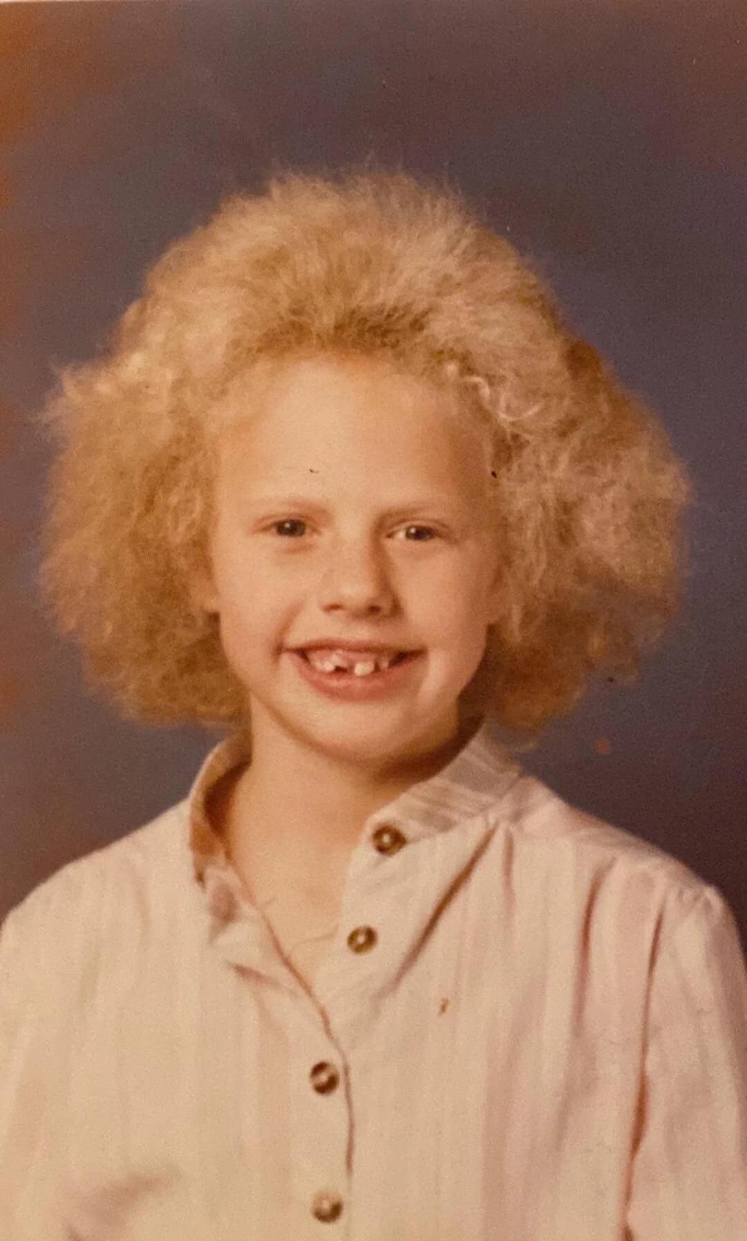 Child with curly blonde hair, missing front teeth, smiling in a school photo.