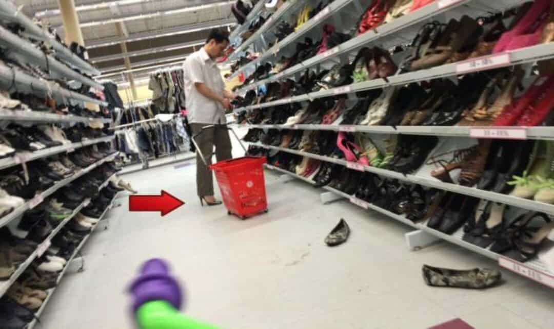 Man shopping in a shoe aisle with a red shopping cart, shoes fallen on the floor.