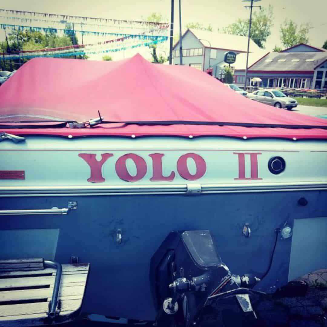 Boat with 'YOLO II' written on it, covered with a pink tarp, parked outside.
