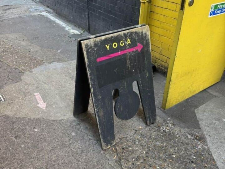 YOGA sign with an arrow pointing right, placed on a sidewalk near a yellow building.