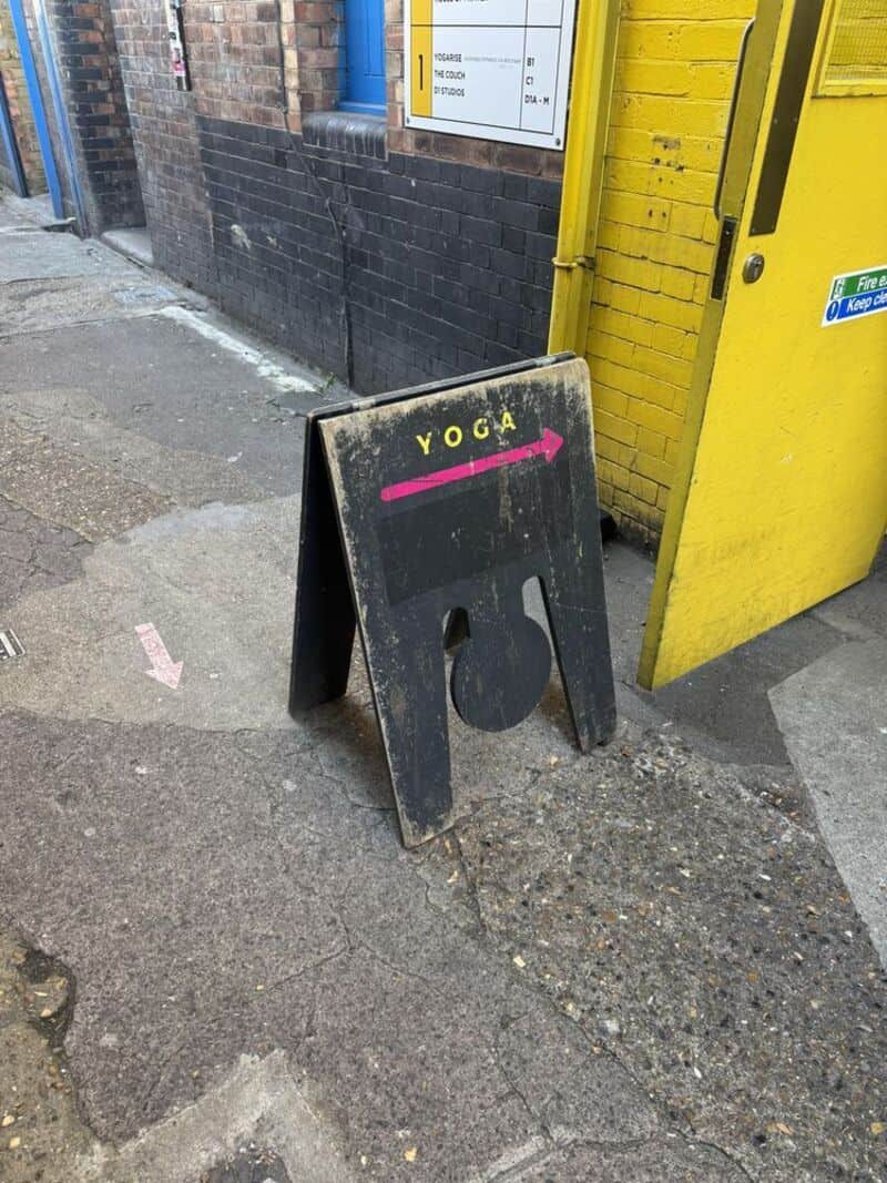 YOGA sign with an arrow pointing right, placed on a sidewalk near a yellow building.