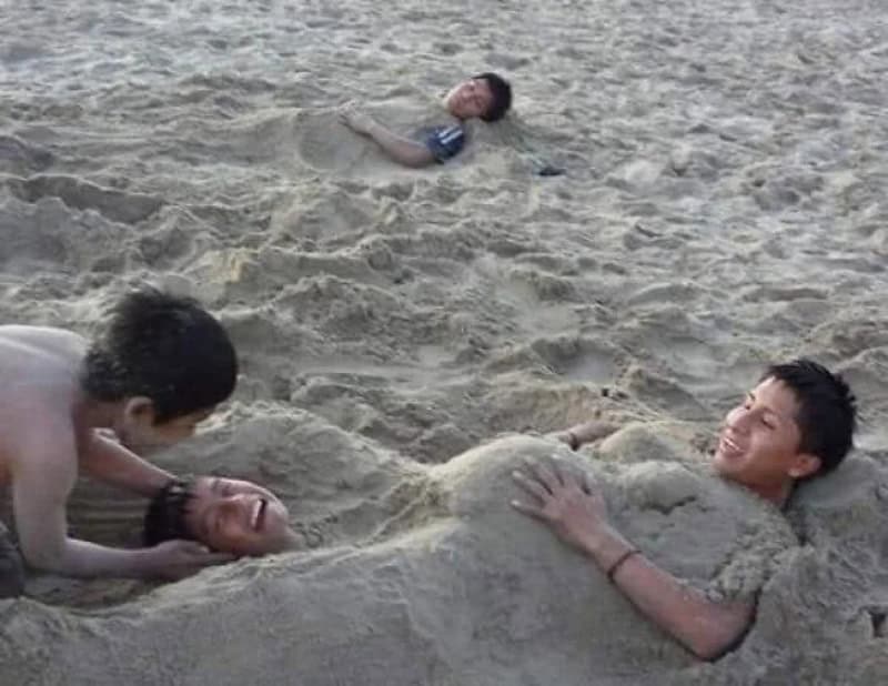 Four boys on a sandy beach, one buried in sand with only head and arms visible.