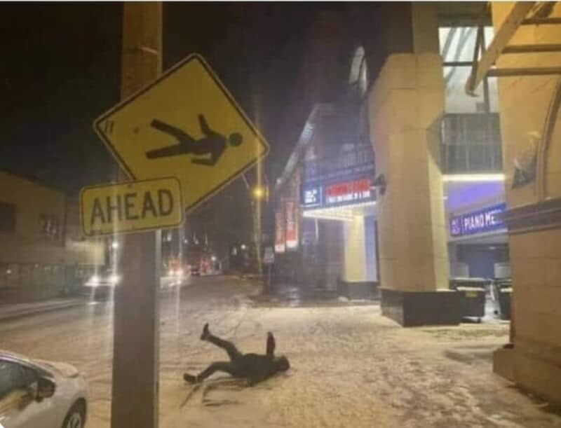 Person lying on the snowy sidewalk near a yellow pedestrian crossing sign that says 'AHEAD'.