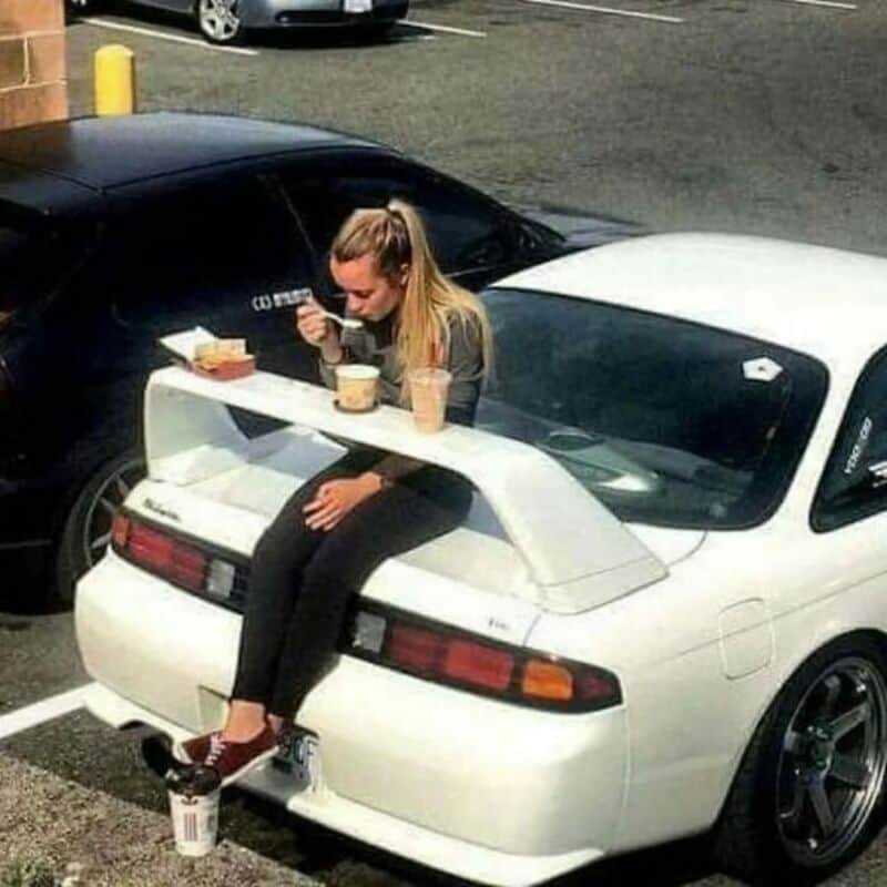 Young woman sitting on a white sports car's rear wing, eating from a takeout container.