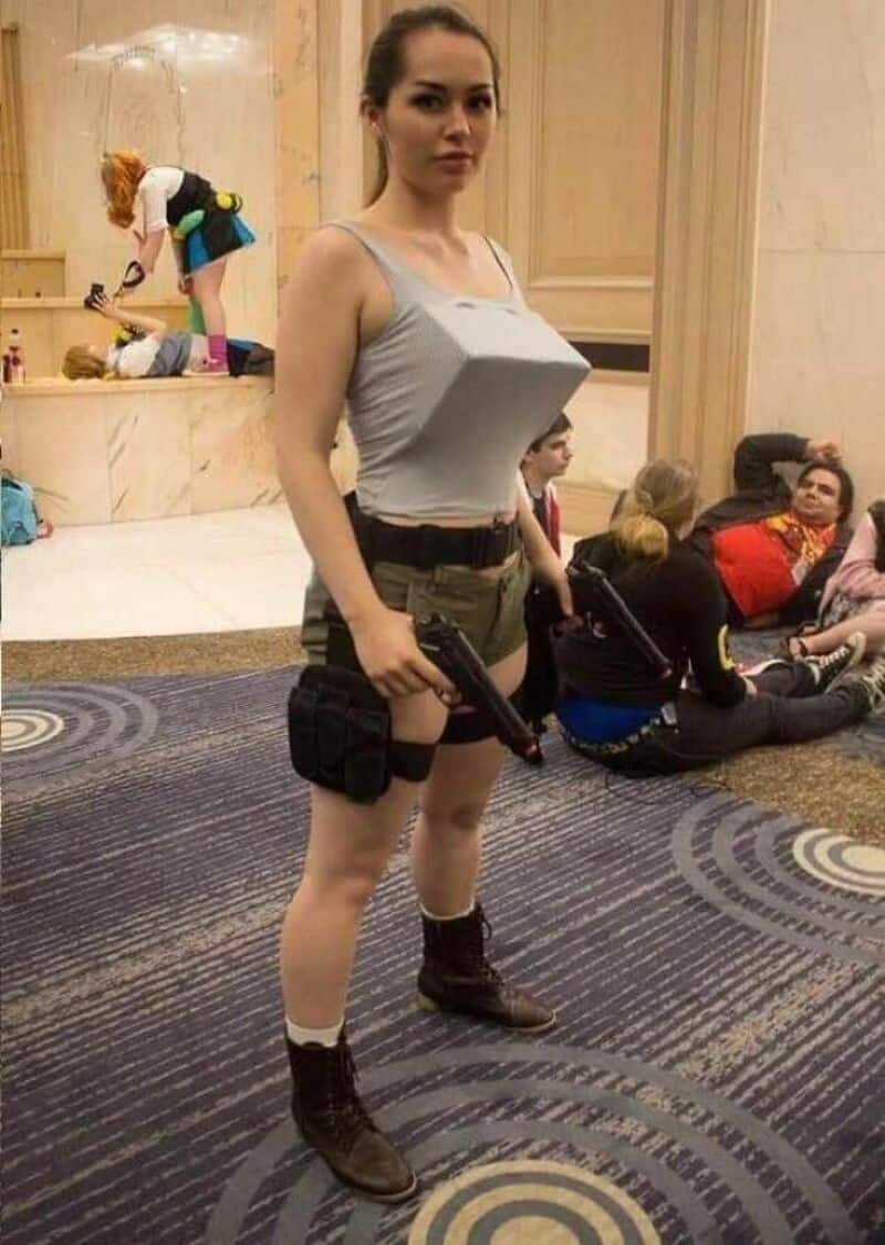 Woman in cosplay costume with a cardboard box on her chest, holding a gun, in a convention setting.