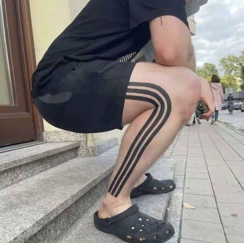 Person squatting on stairs with tattooed leg and black stripes, wearing black Crocs.