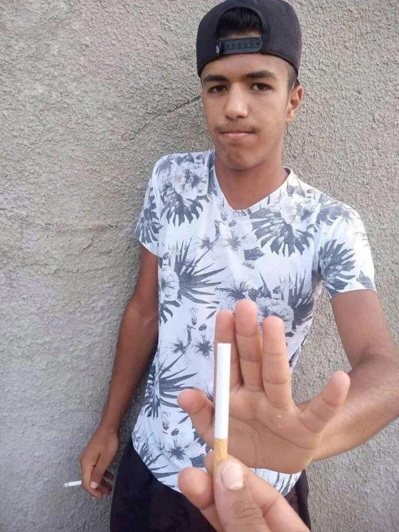 Young man in a floral shirt and black cap holding a cigarette and showing a stop gesture.
