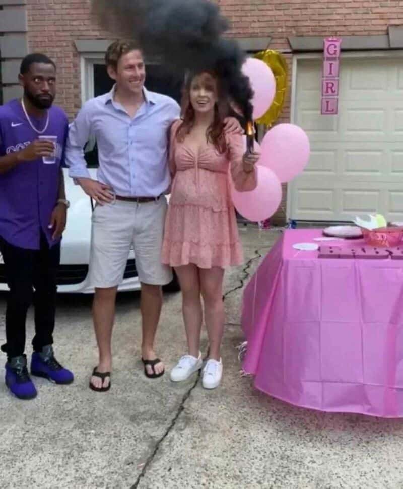 Three friends at a birthday party, one woman holding a smoke bomb, pink balloons, and a pink table.