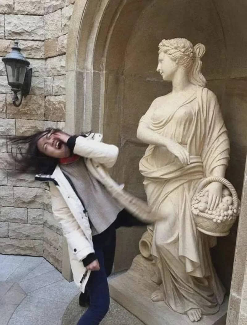 Woman playfully poses next to a classical statue in an archway.