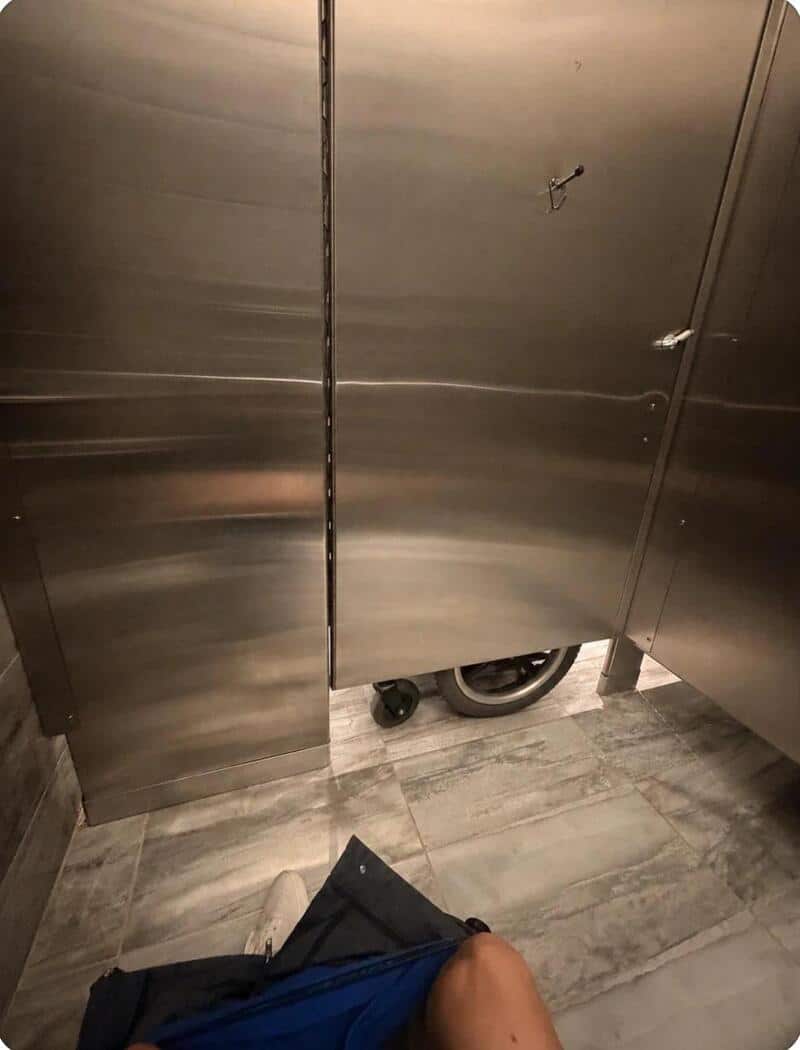 Stainless steel bathroom stall with a gap at the bottom showing a person's knee and a black bag.
