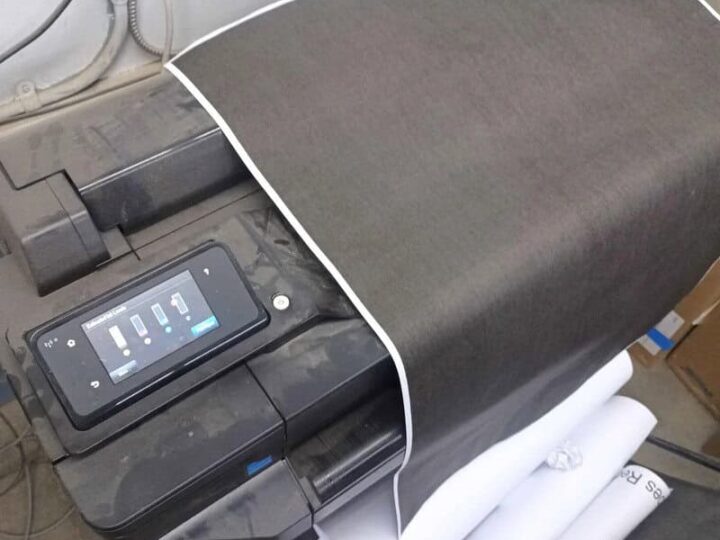 A printer with a large sheet of fabric or paper on it, a can of spray, and rolls of paper underneath.