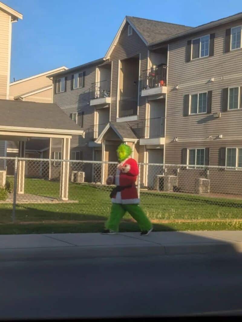 A person dressed as Santa Claus with green hair walking on sidewalk in front of apartment building.
