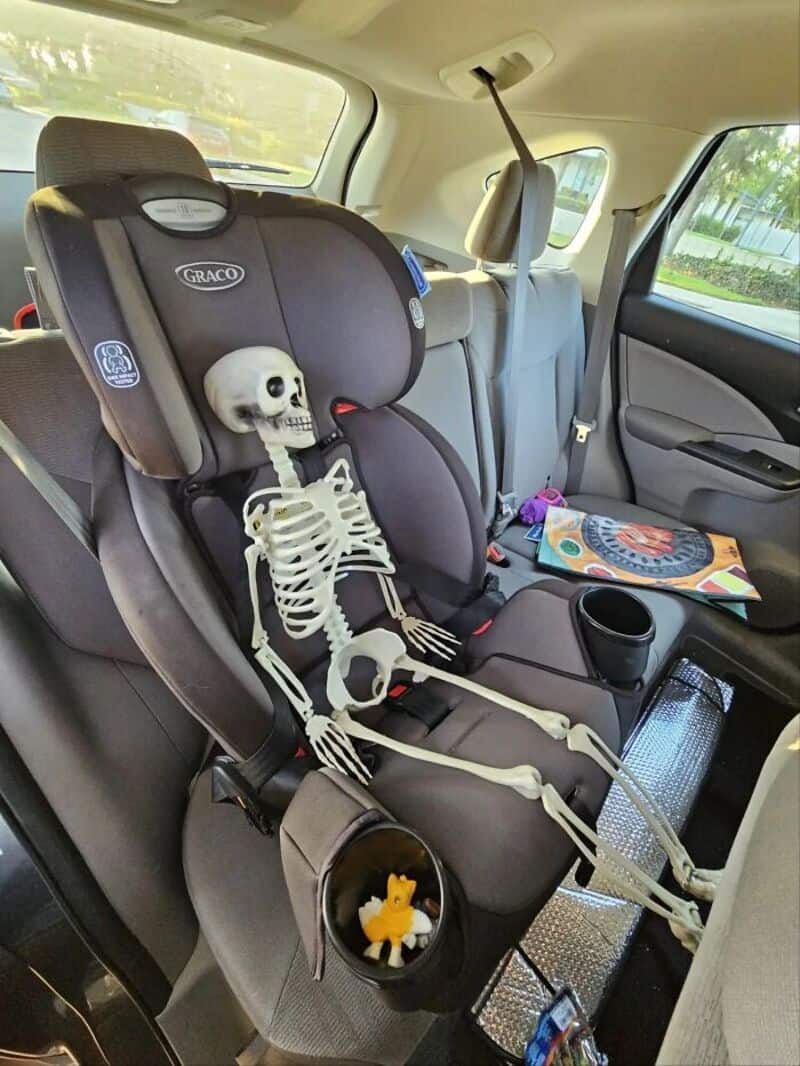 Skeleton toy sitting in a car seat with a rubber duck in a cup holder.
