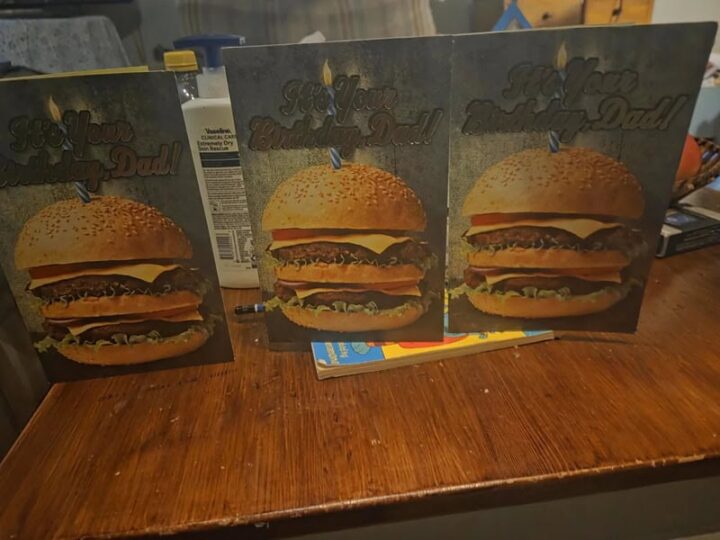 Three birthday cards with burger images and a candle on top, on a wooden table.
