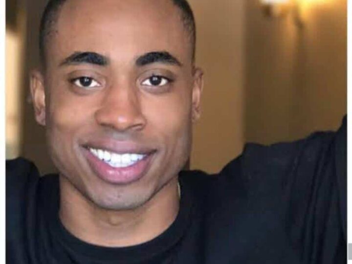 Close-up of a smiling man with a shaved head, wearing a black shirt.