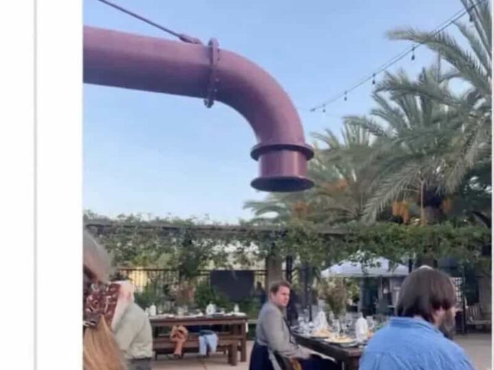 Large rusted pipe hanging above outdoor dining area with people sitting.