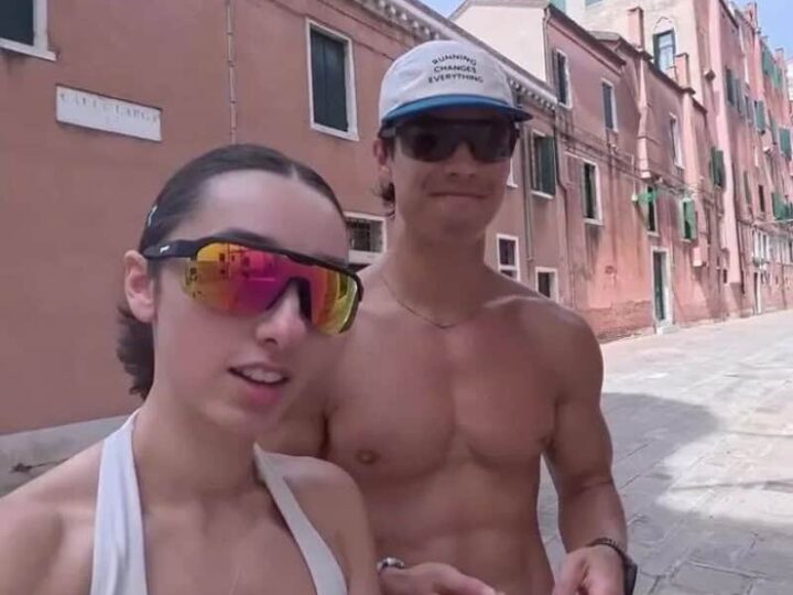 Two tourists, a woman and a man, stand on a street in Venice, Italy, with colorful buildings behind.