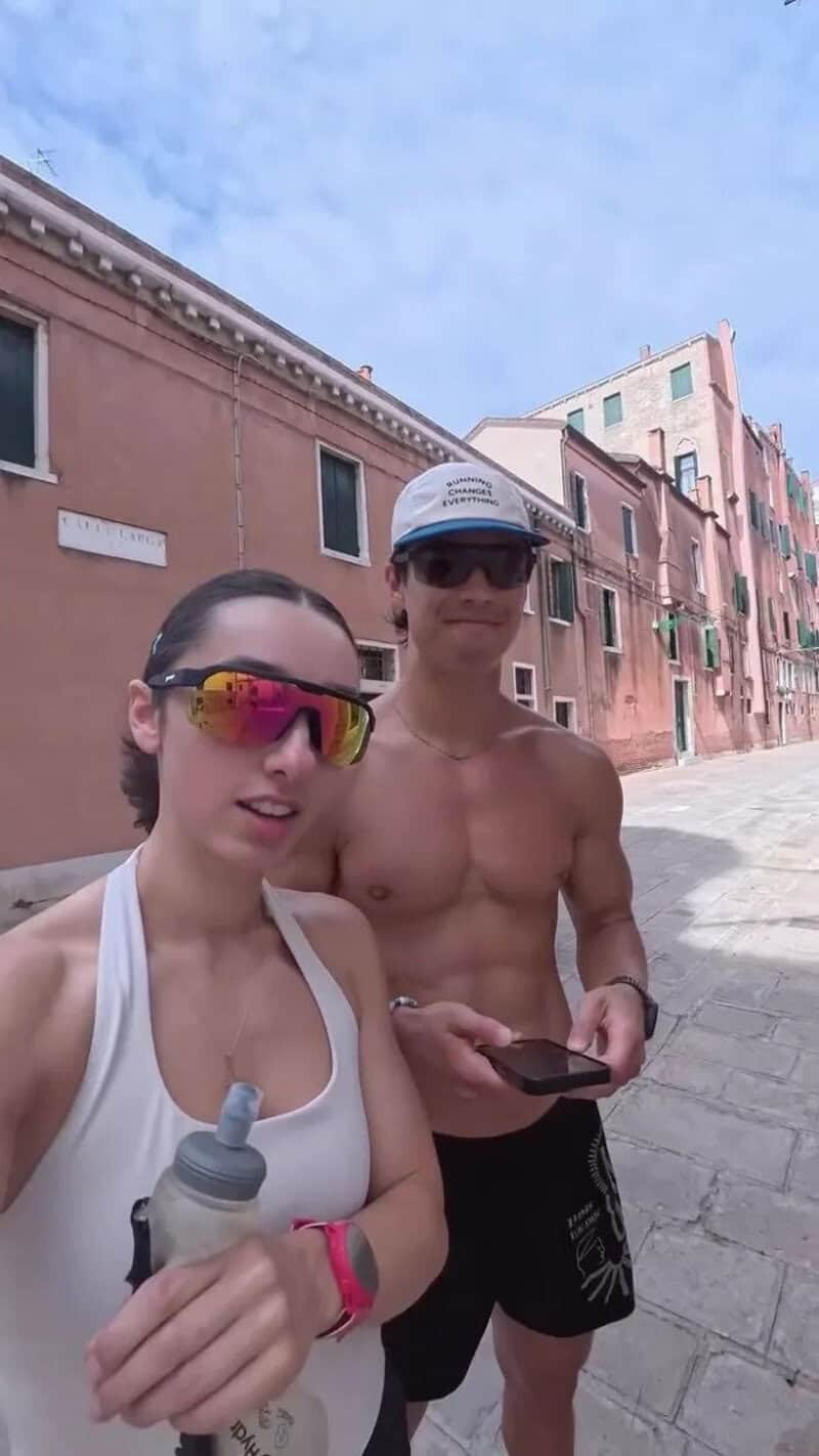 Two tourists, a woman and a man, stand on a street in Venice, Italy, with colorful buildings behind.