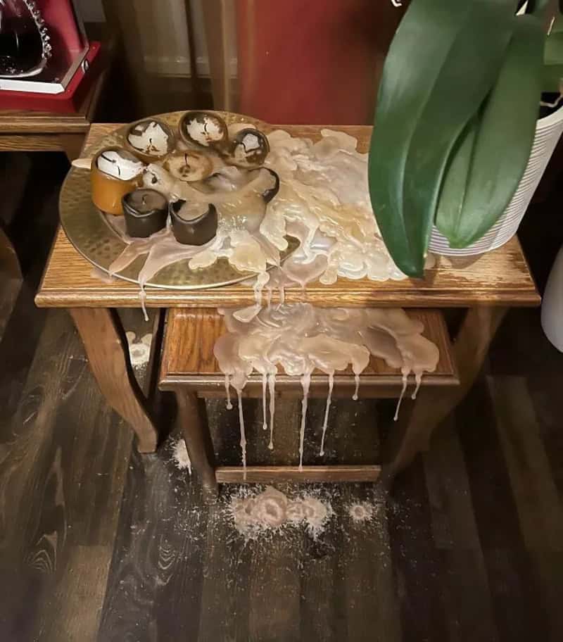 Overflowing candles and wax spilled onto a wooden table and floor, with a plant nearby.