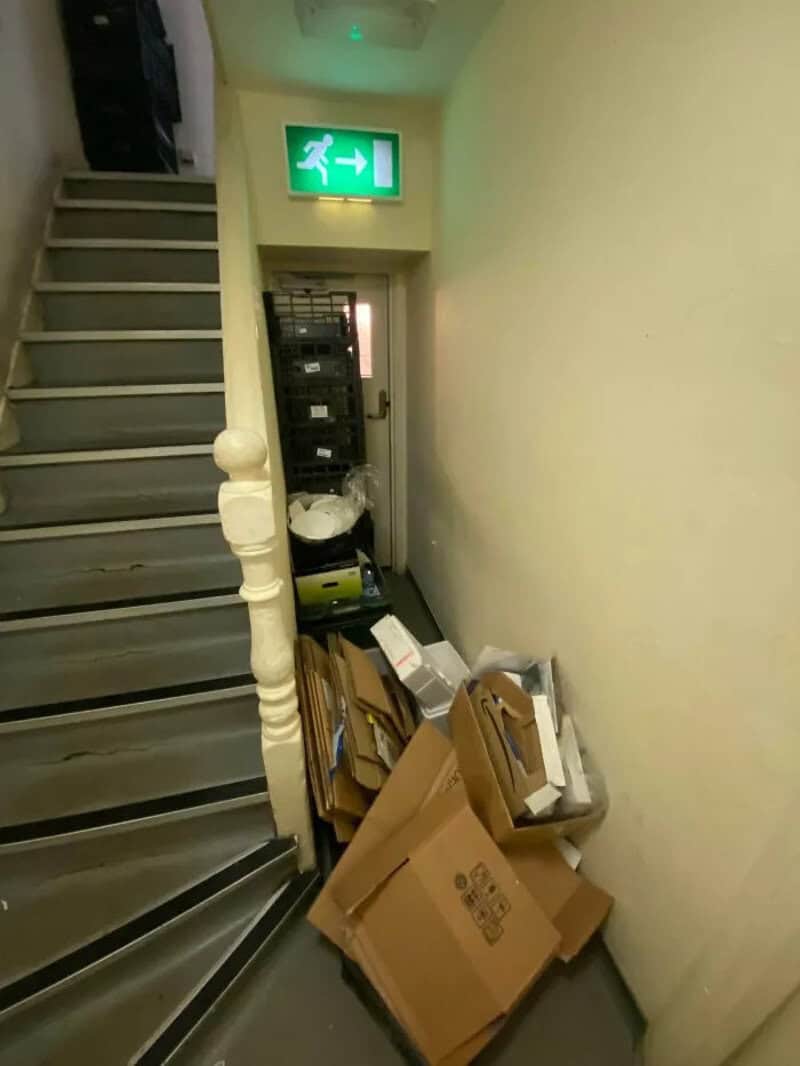 Cluttered corner with boxes and a fire exit sign above a staircase.