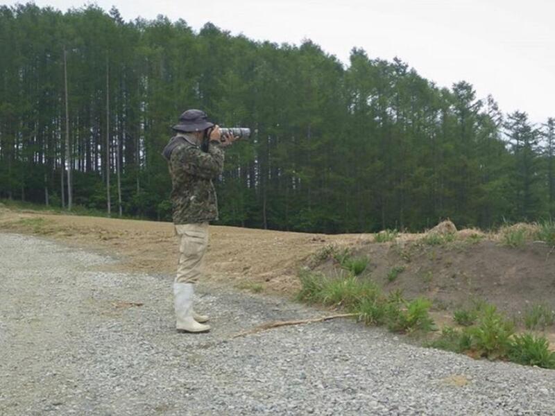 Person in camouflage with a hat using a telescope in a rural area.