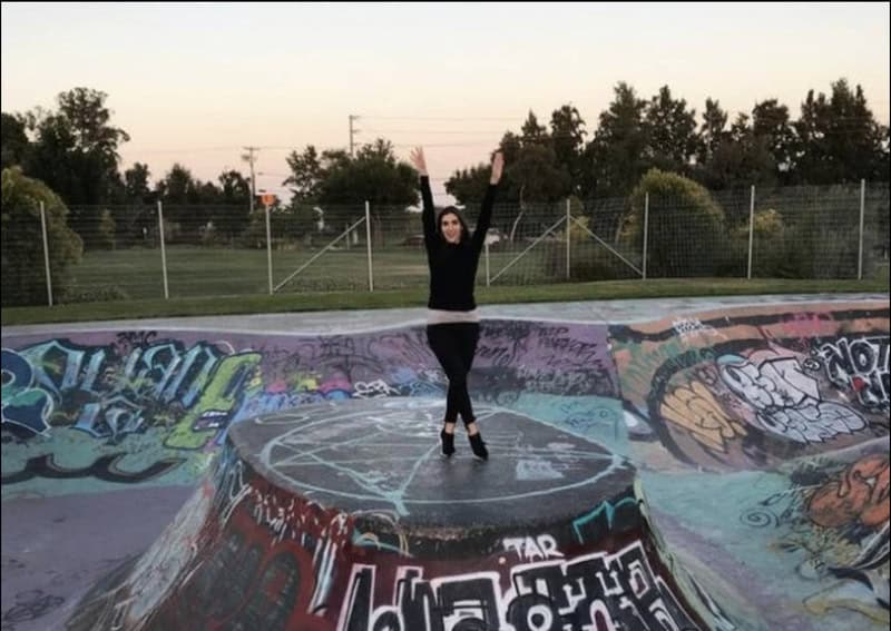 Person standing on a graffiti-covered skatepark with arms raised, sunset in background.