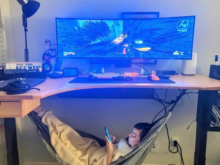 Person relaxing in hammock while playing video game on large monitor.
