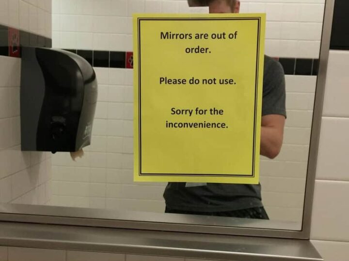 Mirror with a yellow sign that says mirrors are out of order, please do not use, sorry for the incon.