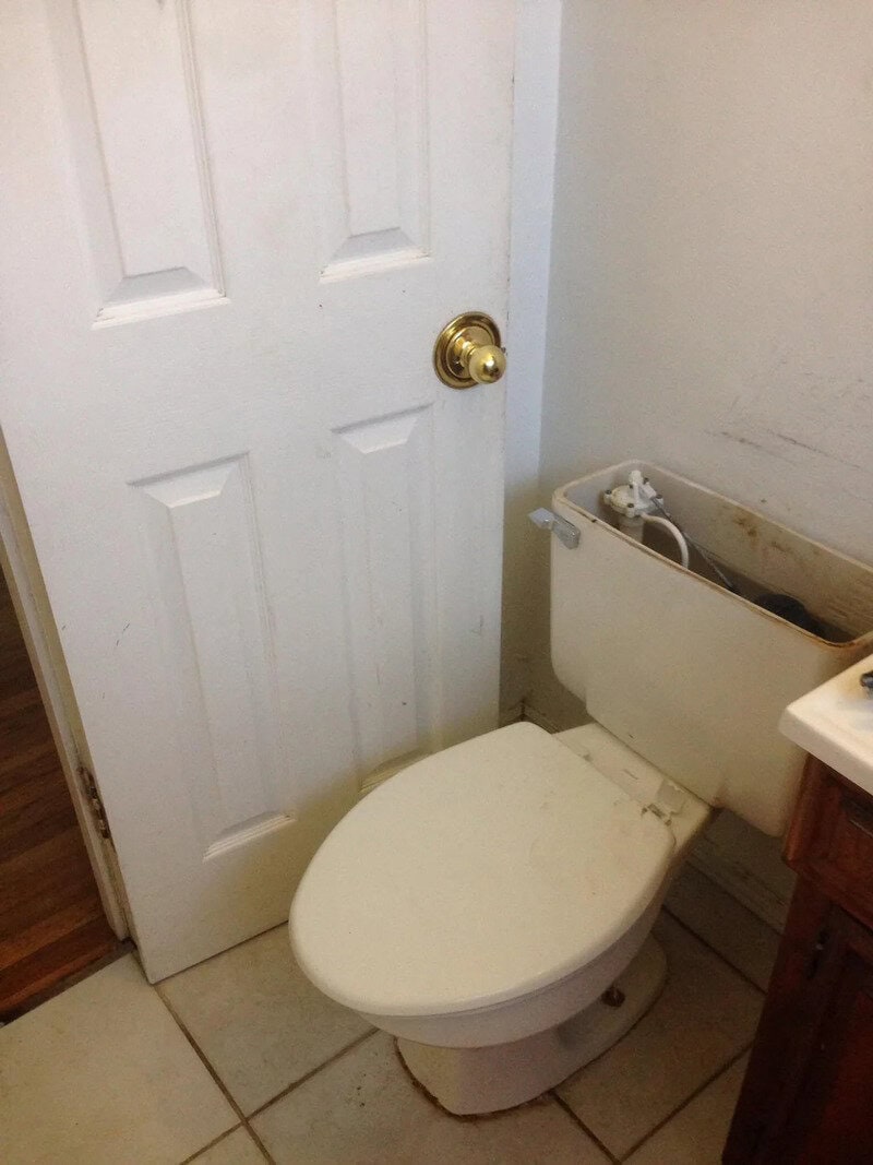 Toilet with missing tank lid and a door behind it in a bathroom.
