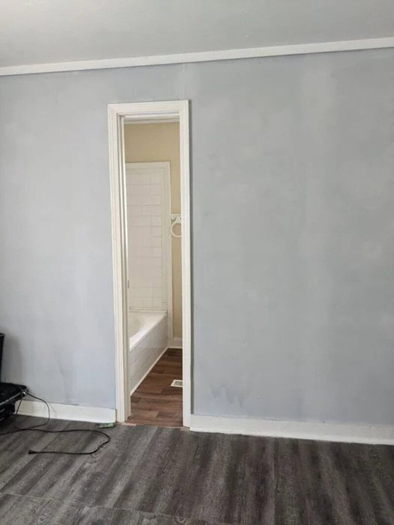 Empty room with a door leading to a bathroom, plain gray walls, and dark wood flooring.