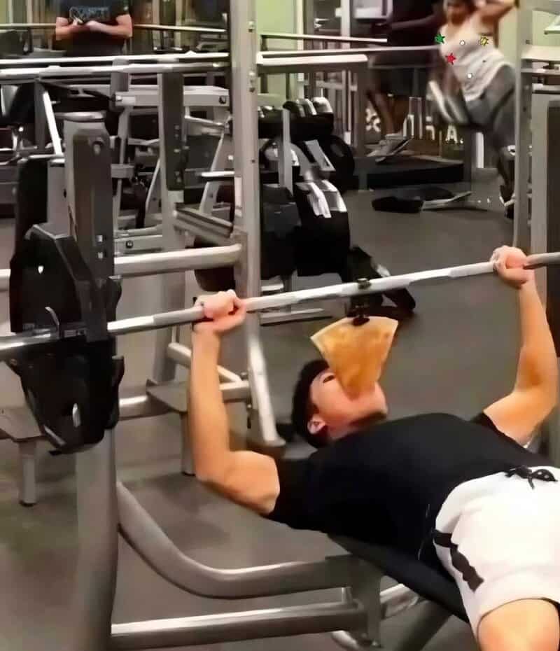 Man lifting a barbell with a slice of pizza in his mouth at the gym.