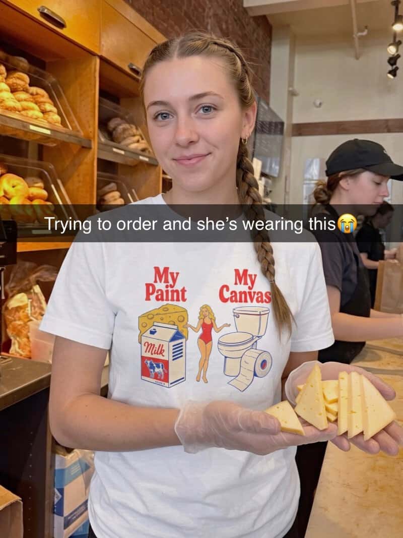 Girl in bakery wearing a T-shirt with a drawing of paint and canvas, holding cheese.