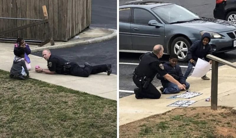 Police officer lying on the sidewalk talking to children, one holding a toy, in a residential area.