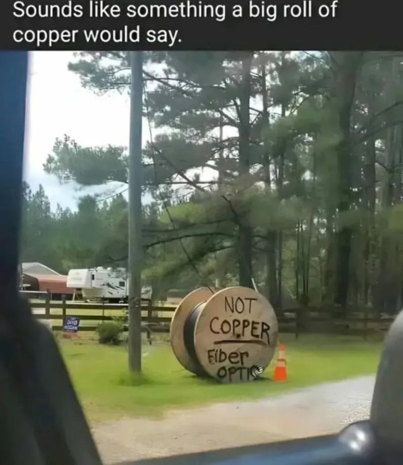 Sign reading 'NOT COPPER Fiber OPTIC' near trees and a road.