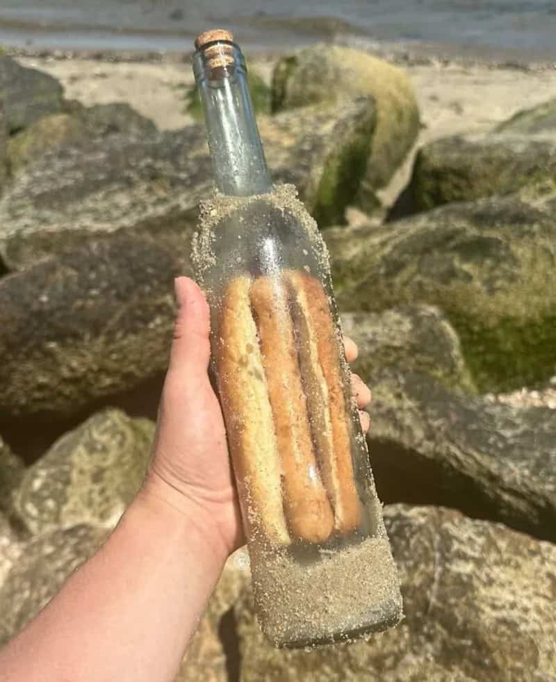 A hand holding a glass bottle with hot dogs inside, on a rocky beach.