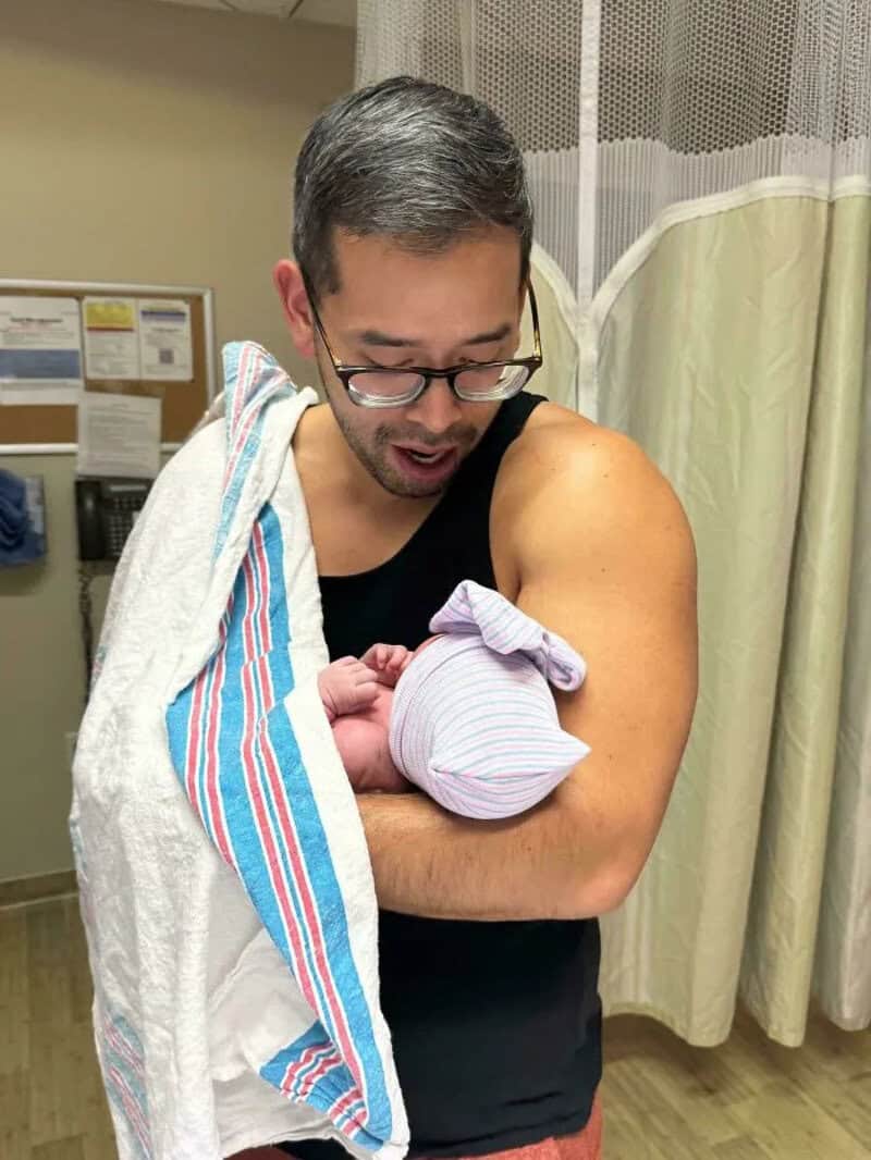 Man holding newborn baby in hospital, looking at baby with love.