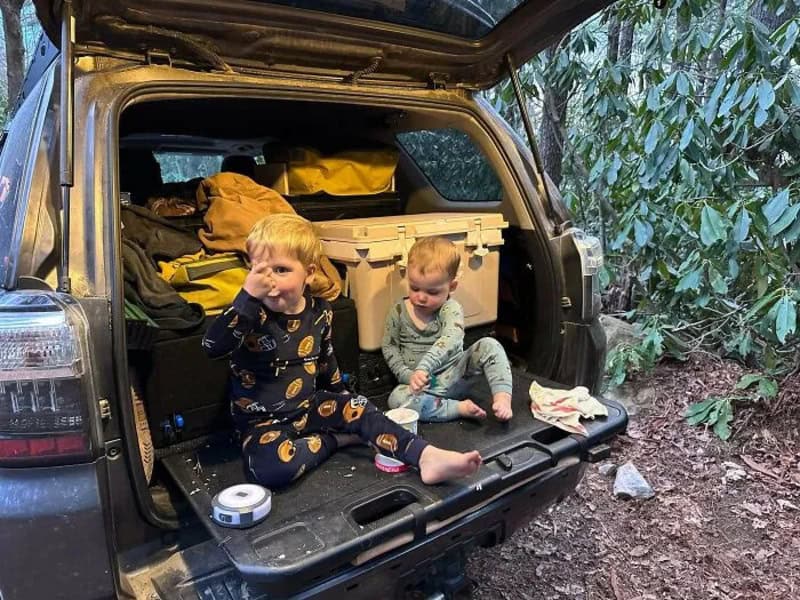 Two young children sitting in the open trunk of a car in a wooded area.