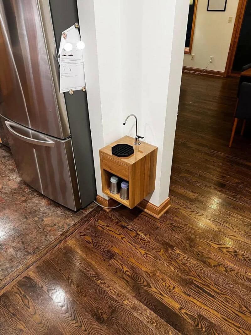 Small wooden shelf with a sink and supplies next to a refrigerator in a kitchen.