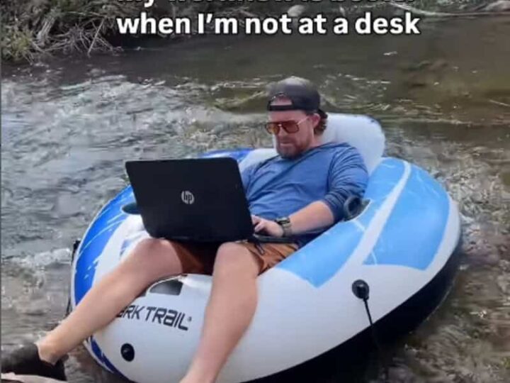 Man working remotely on a laptop in a boat on a river with trees.