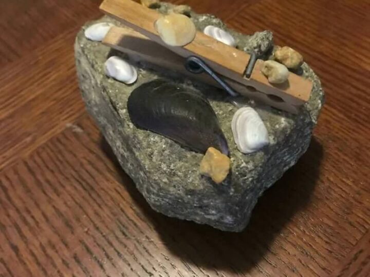 A small rock with a wooden clothespin and tiny stones on top, placed on a wooden surface.