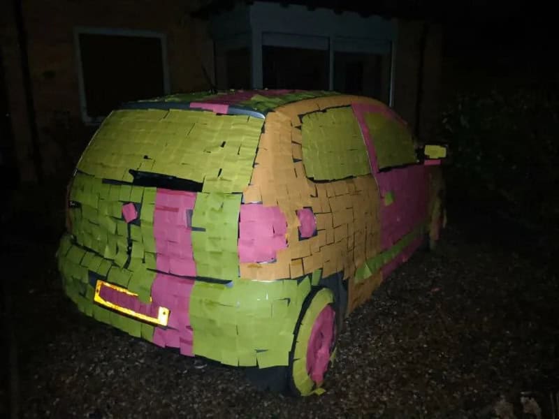 Car covered in colorful sticky notes in a driveway at night.